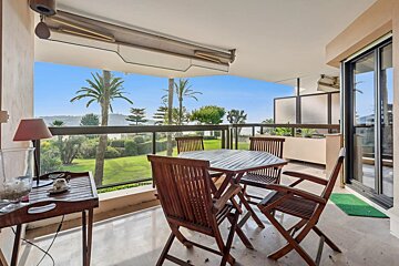 A sunny balcony with wooden table and chairs, overlooking palm trees, a green lawn, and the sea under a clear blue sky.