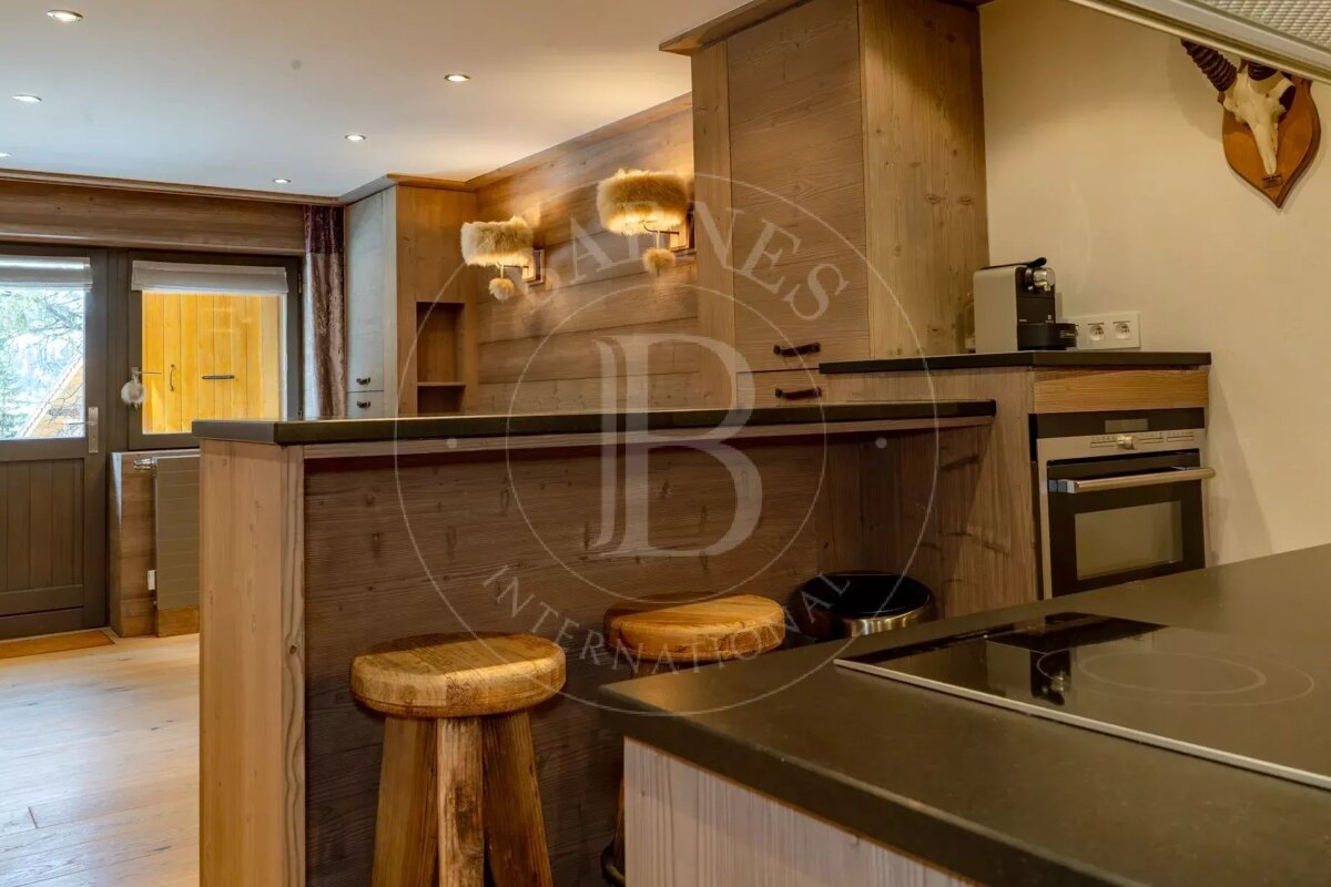 A rustic wooden kitchen with a breakfast bar, stools, and integrated appliances, illuminated by warm lights.