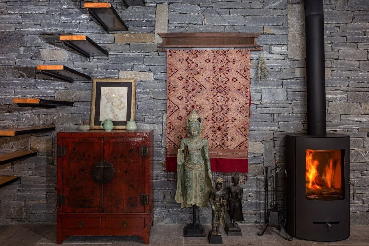 A rustic-modern room features a stone wall, floating stairs, red cabinet with map, Buddha statues, a woven tapestry, and a warm wood-burning stove.