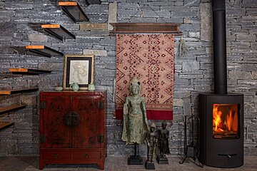A rustic-modern room features a stone wall, floating stairs, red cabinet with map, Buddha statues, a woven tapestry, and a warm wood-burning stove.