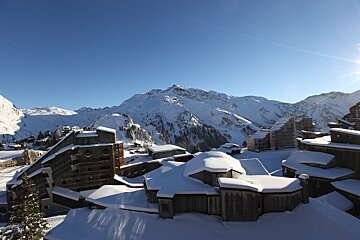 A sunny winter scene of a snow-covered mountain village with unique buildings. Majestic, snow-capped peaks rise under a brilliant blue sky.