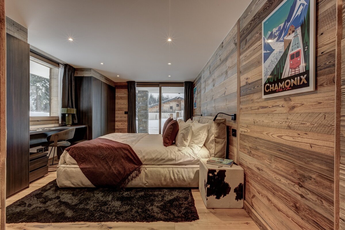 Cozy rustic bedroom with wooden walls, a large bed, and a desk. Features a snowy outdoor view and a vintage Chamonix ski poster.