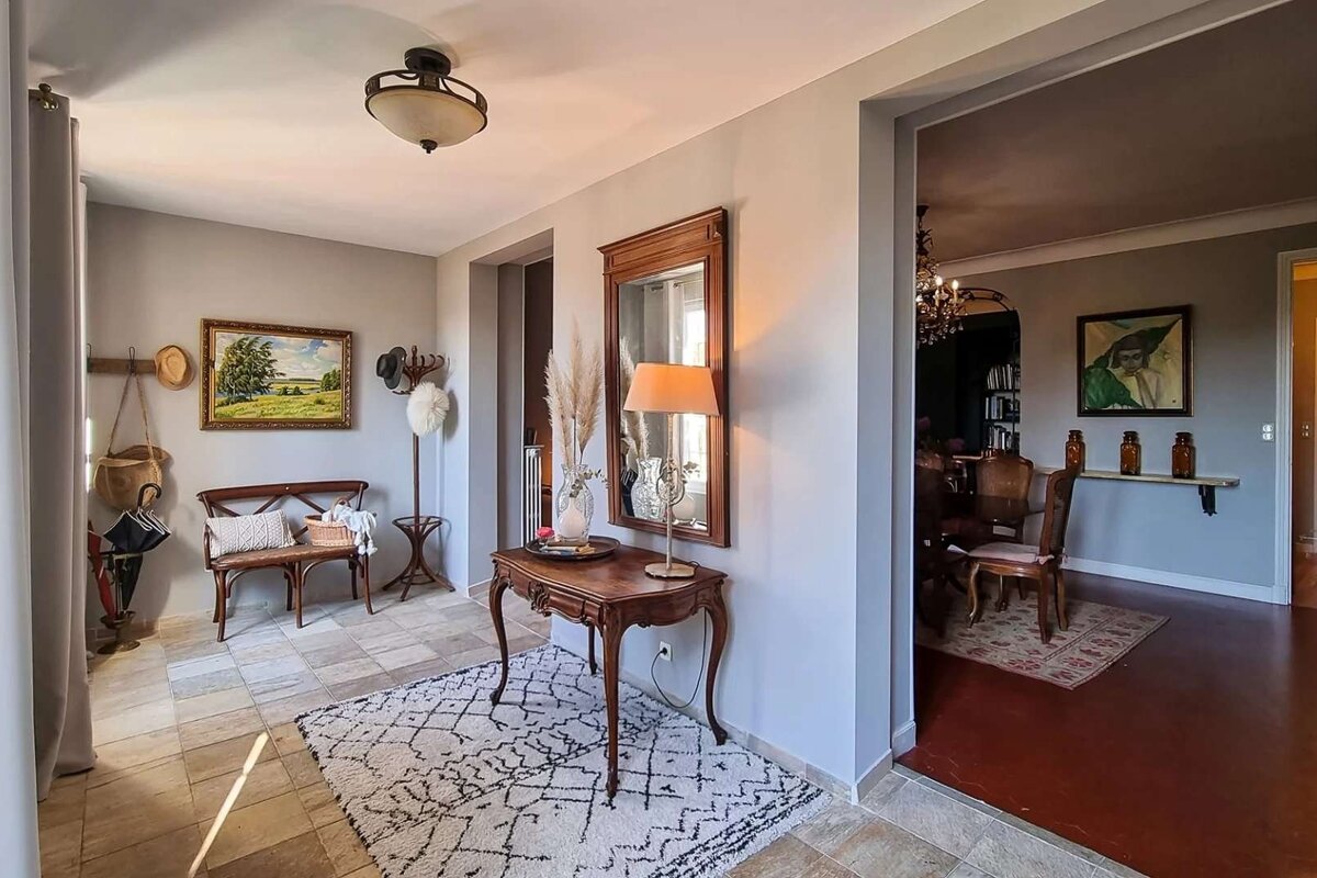 A cozy, light-filled entryway with tiled floors, a patterned rug, wooden bench, and console table with a lamp and mirror. A landscape painting hangs on the wall.
