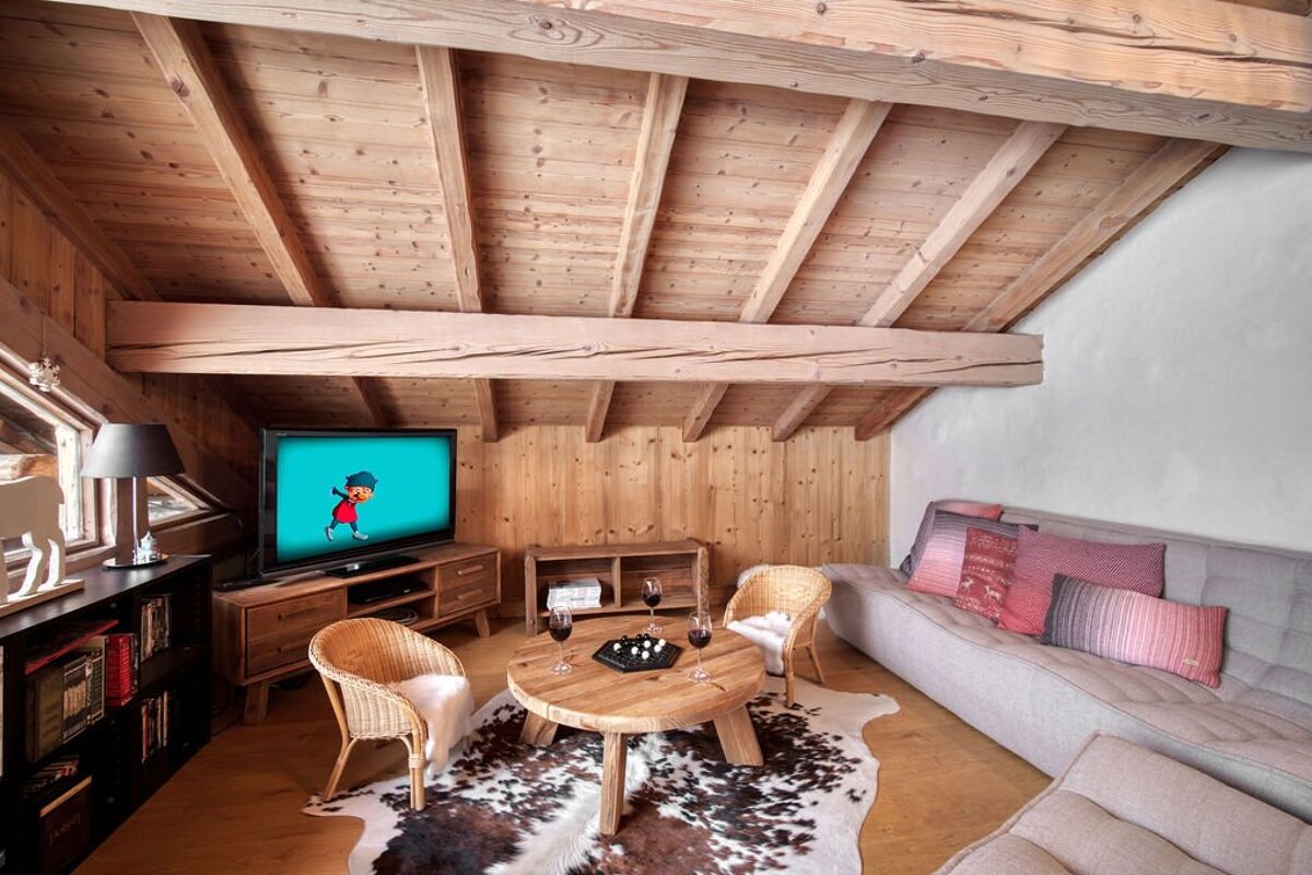 Rustic chalet loft featuring wooden beamed ceilings, a gray sofa, wicker chairs, a cowhide rug, and a TV.
