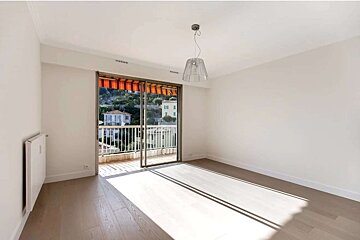 Bright, empty room with white walls, wooden floor, and a sliding glass door to a balcony. Sunlight streams in, revealing a view of houses and hills.