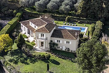 Aerial view of a sprawling villa with a red-tiled roof, swimming pool, and lush green gardens surrounded by olive trees.