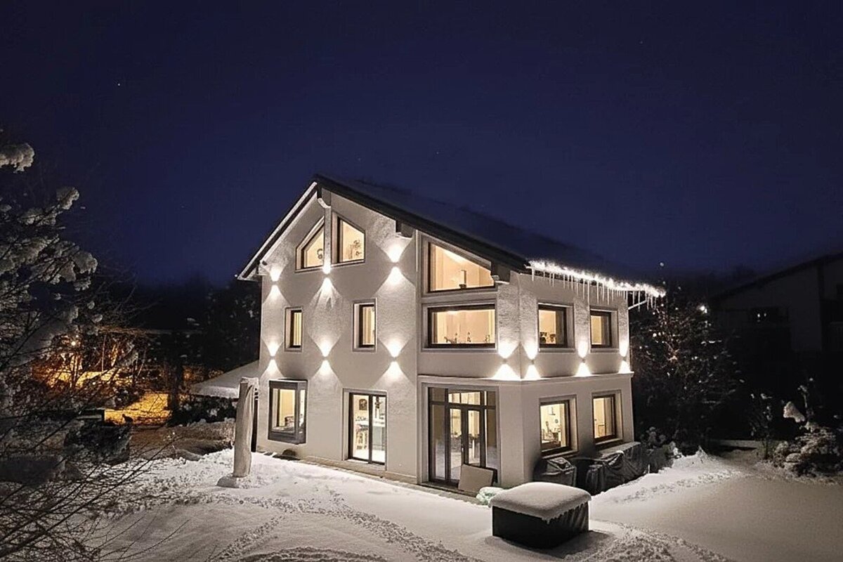 A modern, brightly lit house stands in a snowy landscape at night. Warm lights illuminate its walls, and icicles hang from the roof.