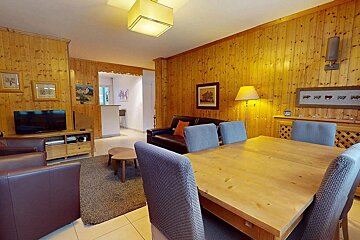 A warm, wood-paneled living and dining room with a large wooden table, chairs, sofas, and a TV, creating a cozy chalet atmosphere.