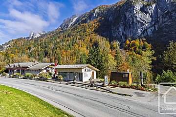 A mountain road winds past buildings with an autumn-colored forest and rocky cliffs in the background under a bright blue sky.