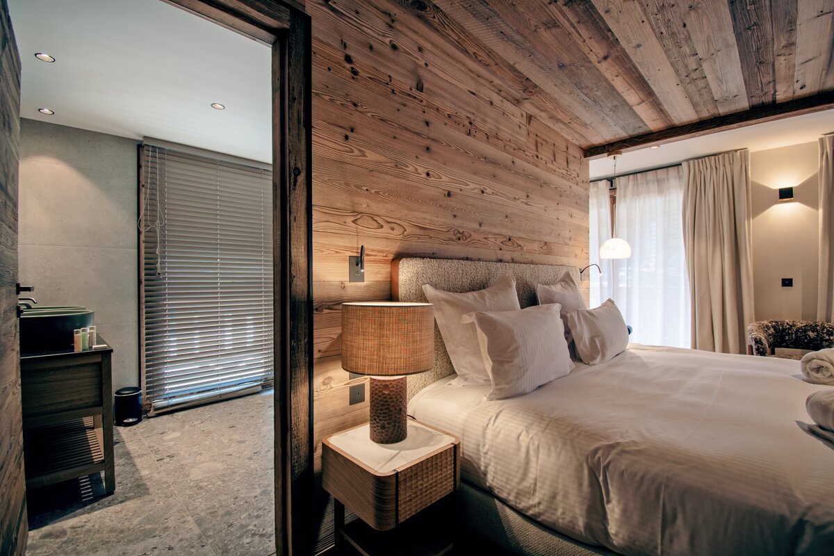 Cozy, rustic-modern chalet bedroom with warm wood paneling, a large bed, and an adjacent tiled bathroom visible through an open door.