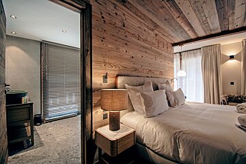 Cozy, rustic-modern chalet bedroom with warm wood paneling, a large bed, and an adjacent tiled bathroom visible through an open door.