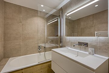 Modern bathroom featuring beige tiled walls, a bathtub with wooden trim, a glass divider, and a sleek white vanity with a backlit mirror.