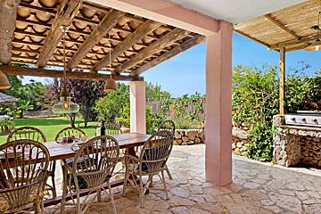 Rustic outdoor patio with dining table, wicker chairs, BBQ, and lush garden view under a wooden beamed ceiling.