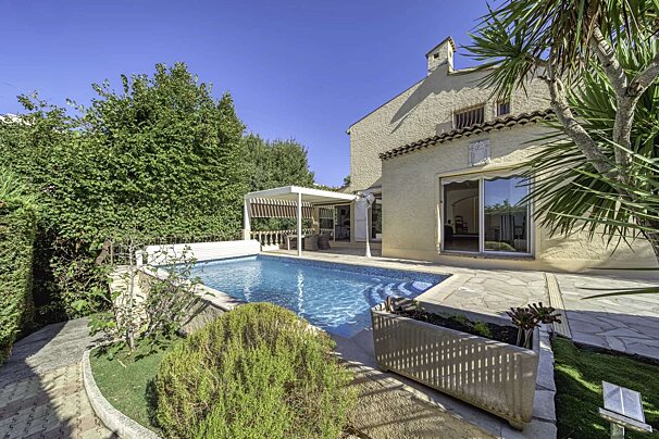 Sunny Mediterranean-style house with a blue swimming pool, patio, and lush garden under a clear sky.