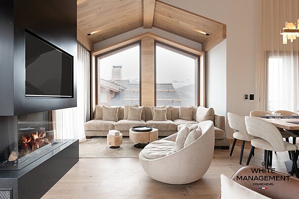 A chic mountain chalet living room with a fireplace, panoramic windows showcasing snowy peaks, a neutral sectional sofa, and modern seating.