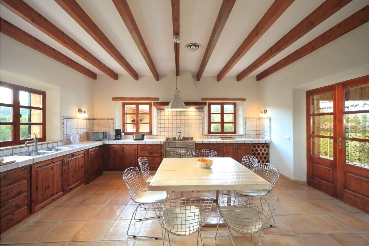 Spacious rustic kitchen with exposed wooden ceiling beams, terracotta floor, and white tiled counters. A large dining table with modern wire chairs sits centrally.