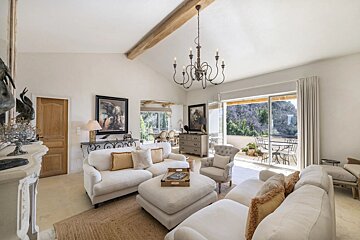 A bright, elegant living room with white sofas, a large chandelier, vaulted ceiling, and glass doors opening to a scenic balcony.