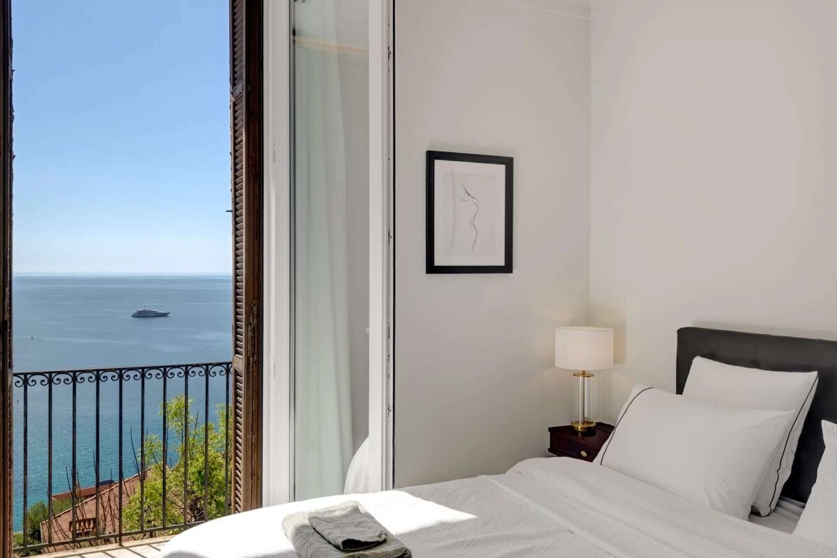 A bright bedroom with an open balcony overlooking a beautiful blue sea with a yacht. Features a white bed and minimalist decor.