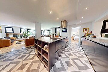 Modern open-plan kitchen and living area with a large island, geometric floor tiles, and a view into a bedroom.