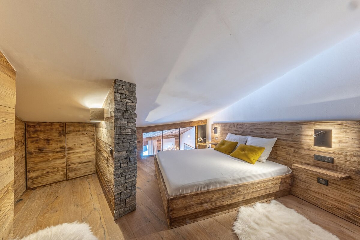Cozy, rustic attic bedroom featuring a wooden bed, walls, and a stone accent partition. Yellow pillows add a pop of color.