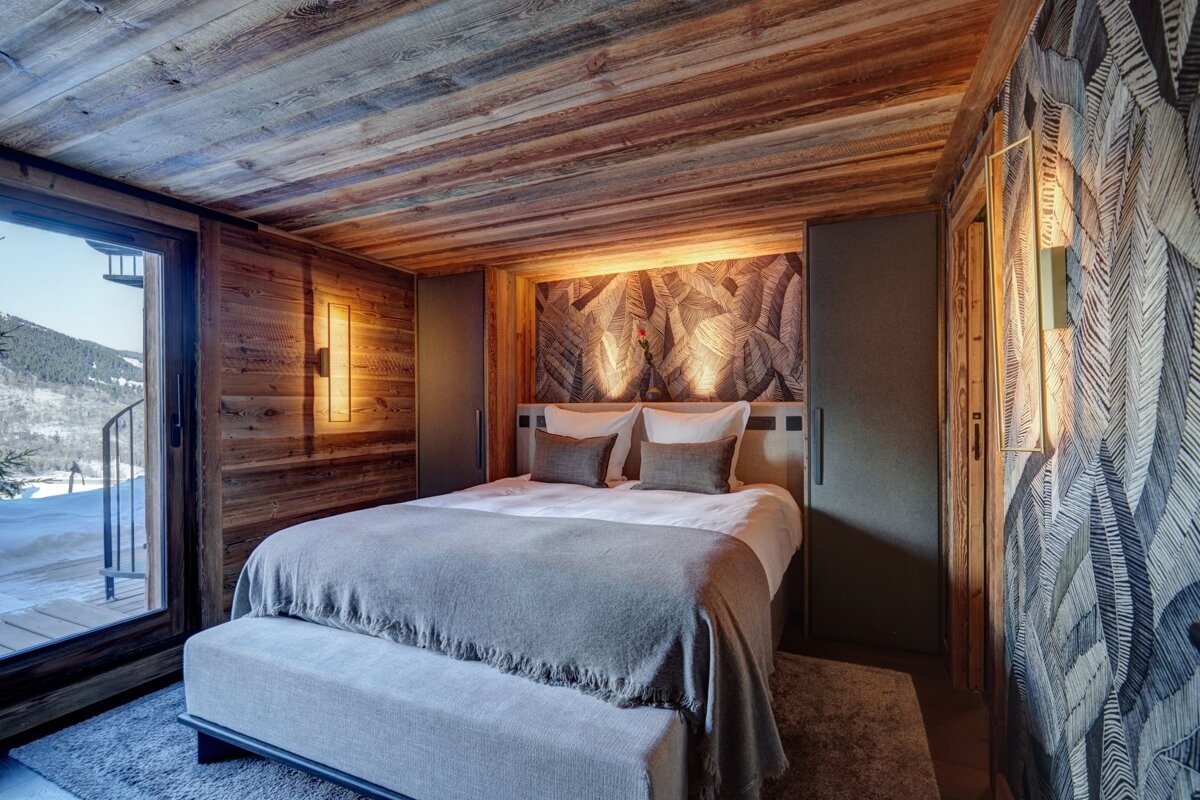 Cozy, wood-paneled bedroom with a plush bed, nature-patterned wallpaper, and a large window offering a snowy mountain view. Warm lighting enhances the ambiance.