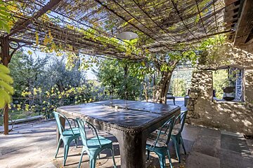 A sun-dappled outdoor patio with a vine-covered pergola, a large dark metal table, and teal chairs. Lush greenery surrounds the rustic stone building.