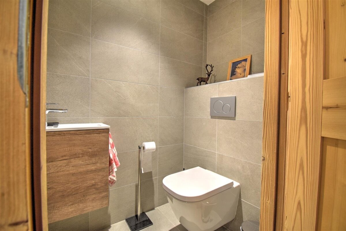 A modern, grey-tiled toilet room featuring a white toilet, a wooden vanity with a sink, and a decorative shelf. Pine wood frames the entrance.