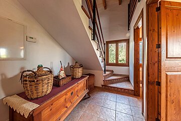 A warm, rustic hallway with a wooden staircase, large chest, woven carboys, and a window looking out to trees. Tiled floor.
