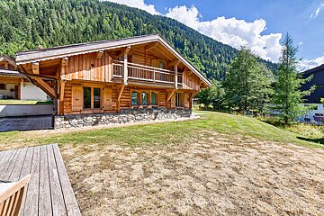 A large wooden house with a balcony sits in the middle of a lush green forest