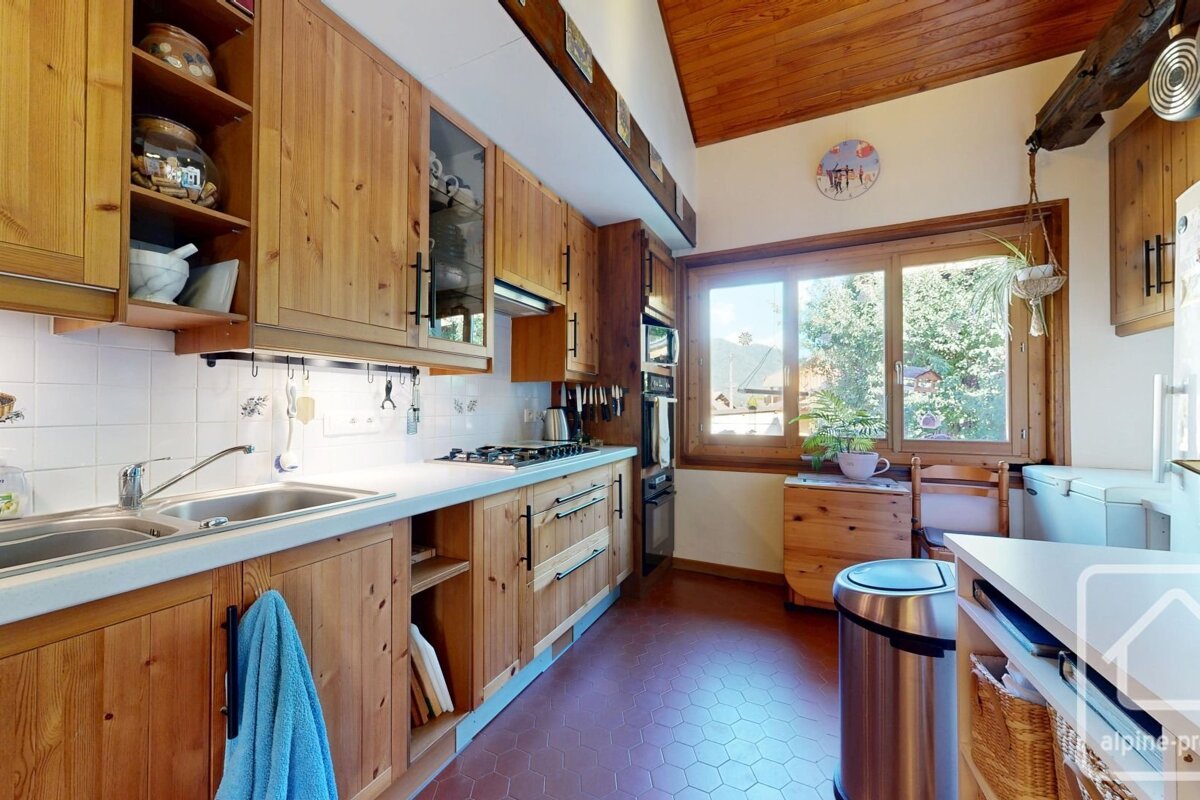 Cozy, rustic kitchen with natural wood cabinets, white counters, and red hexagonal tiled floor. A large window overlooks green trees.
