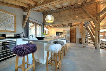 A cozy, rustic-modern chalet kitchen features exposed wooden beams, a sleek island with colorful fluffy stools, and an open layout with bright windows.
