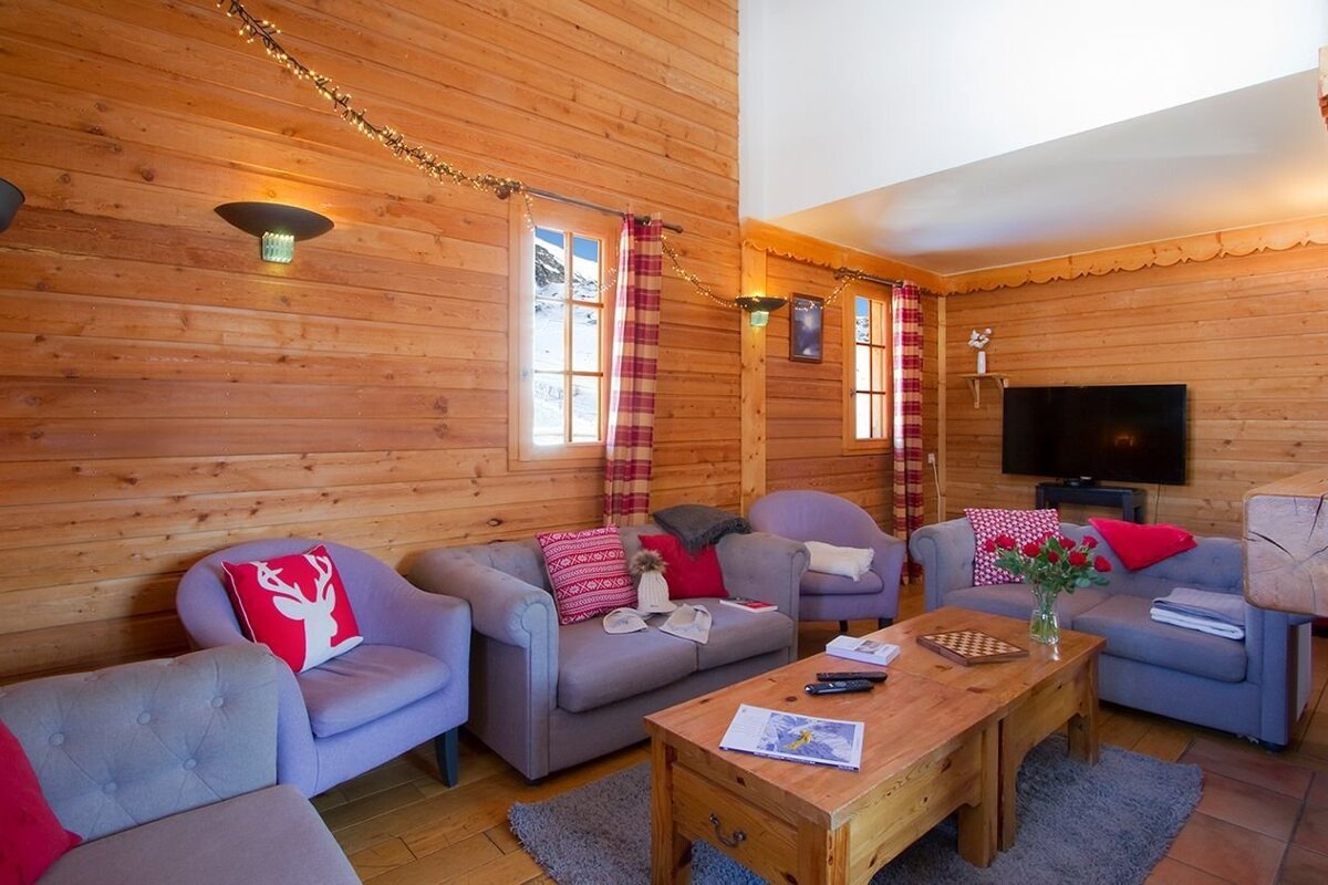 A cozy, wood-paneled cabin living room with gray and purple seating, red accents, string lights, and windows looking out to snowy mountains.
