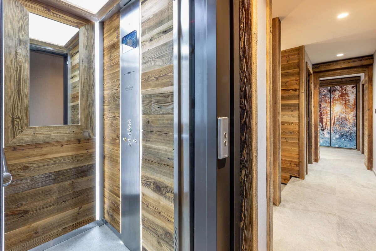 A modern elevator with rustic wood and steel accents opens to a hallway featuring wood paneling and a snowy forest mural with a sunset glow.
