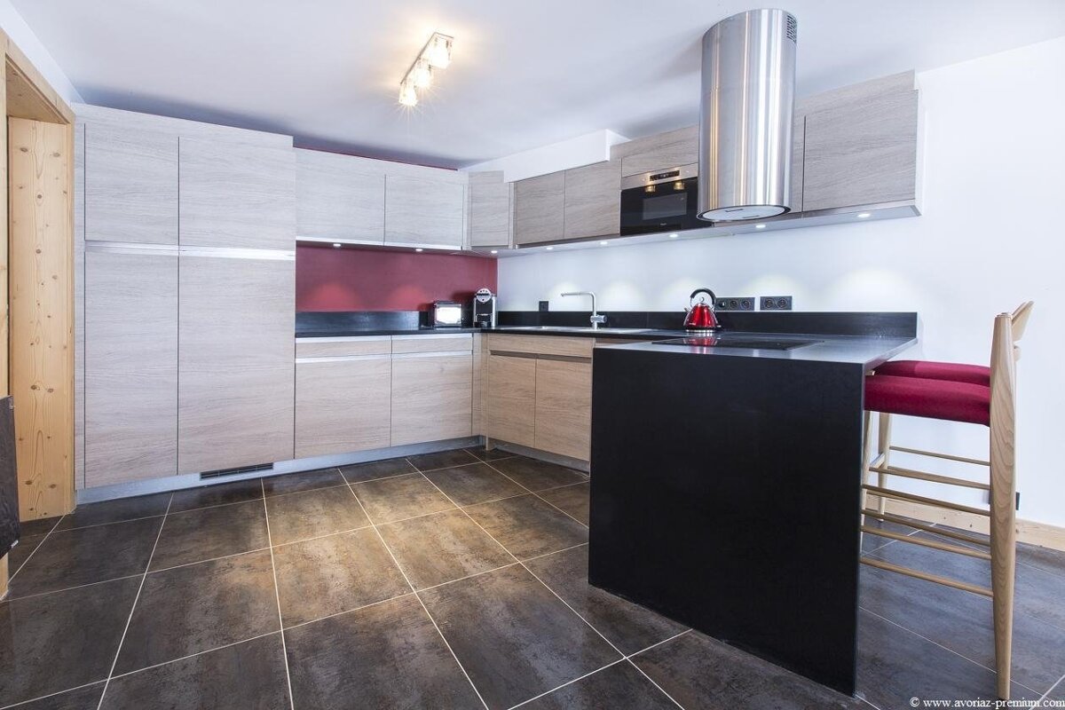 Modern kitchen with light wood cabinets, dark tile floor, black island, red bar stool, and a red accent wall.