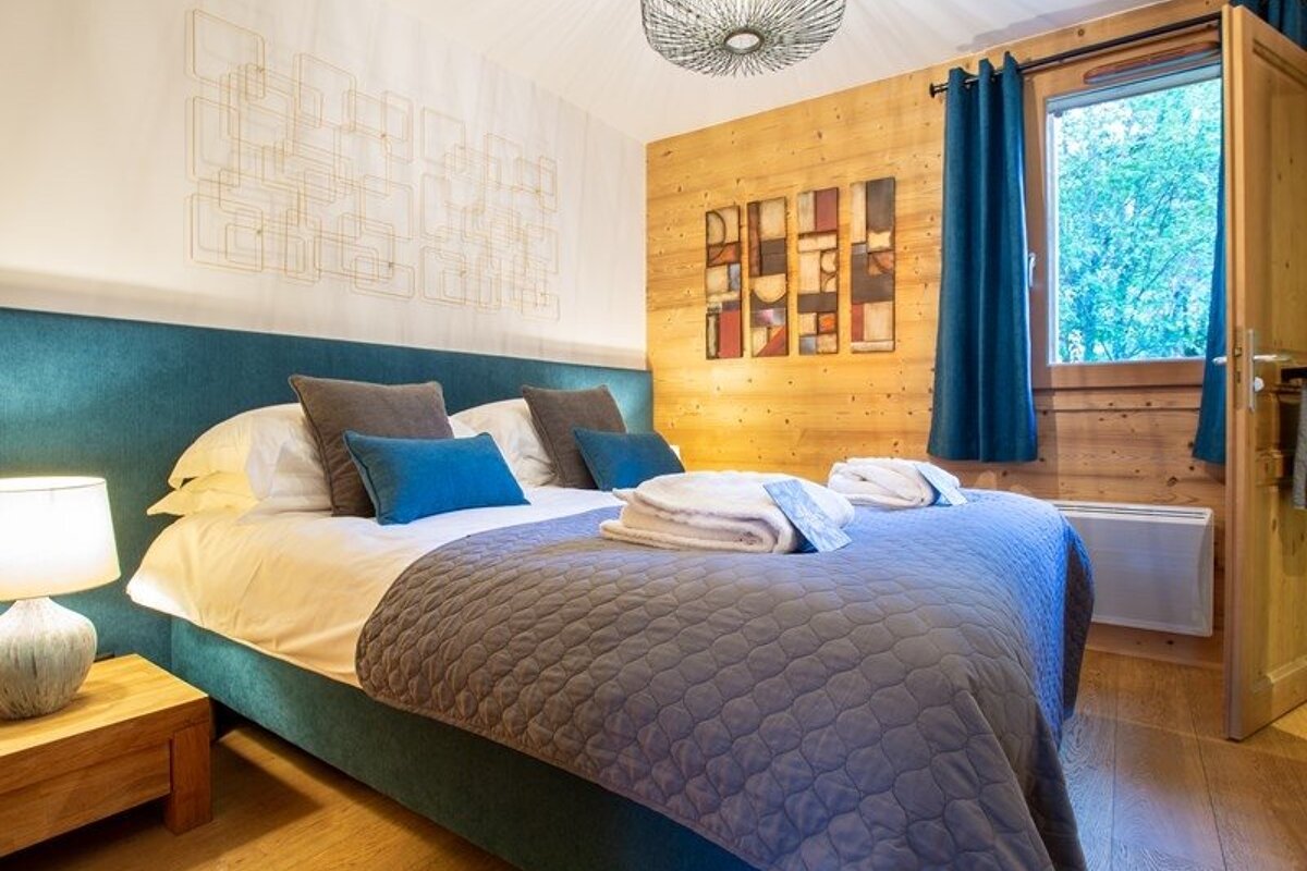 Cozy, modern bedroom featuring a teal upholstered bed, rustic wood-paneled walls, geometric decor, and a window with a blue curtain.