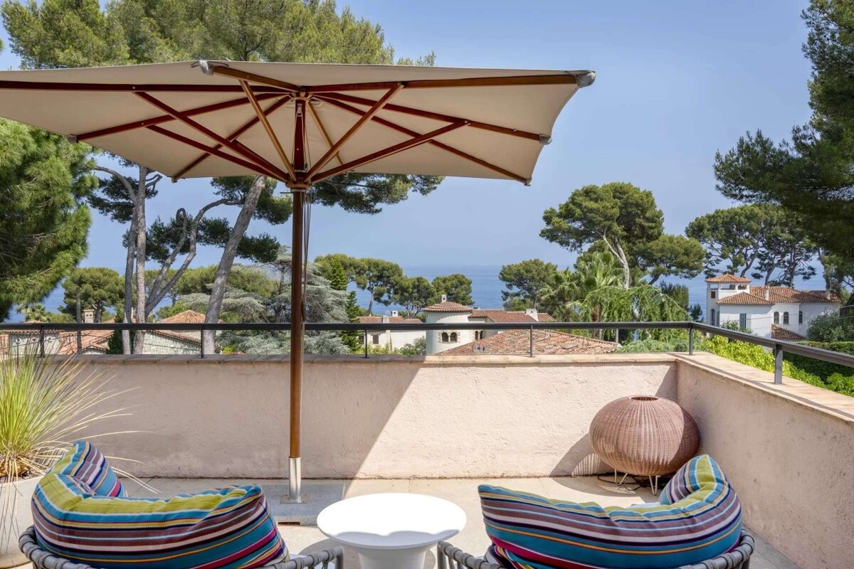 Sunny balcony overlooking the sea and coastal homes, with a large umbrella and colorful striped chairs for relaxation.