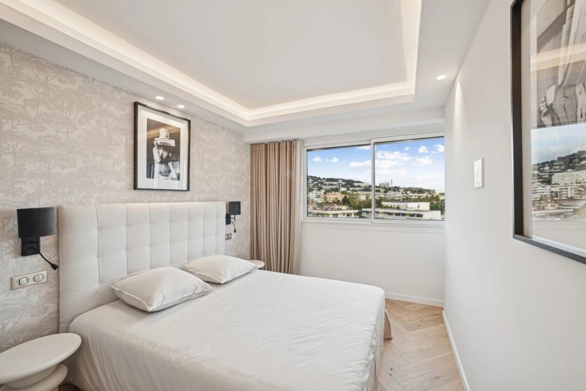 Bright, modern bedroom featuring a white bed, patterned wallpaper, recessed lighting, and a large window overlooking a city landscape.