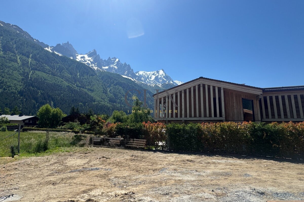 A wooden chalet sits before a backdrop of snow-capped mountains and green slopes under a clear blue sky.