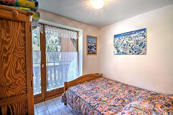 A cozy bedroom with a wooden bed, colorful patterned duvet, and glass balcony doors overlooking a green forest.
