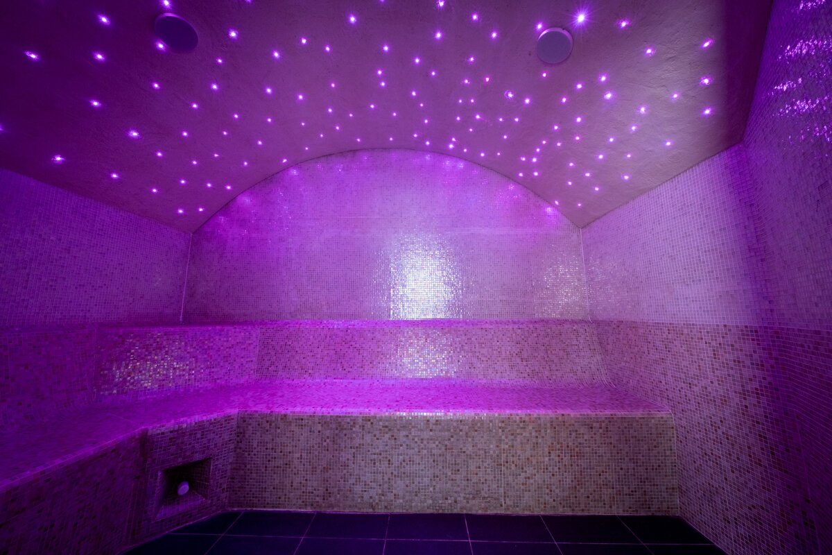 A purple-lit steam room with a curved, star-light ceiling. Tiled walls and benches create a serene and mystical spa ambiance.