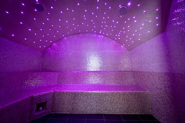 A purple-lit steam room with an arched ceiling, dotted with star-like lights, and mosaic-tiled walls and benches.