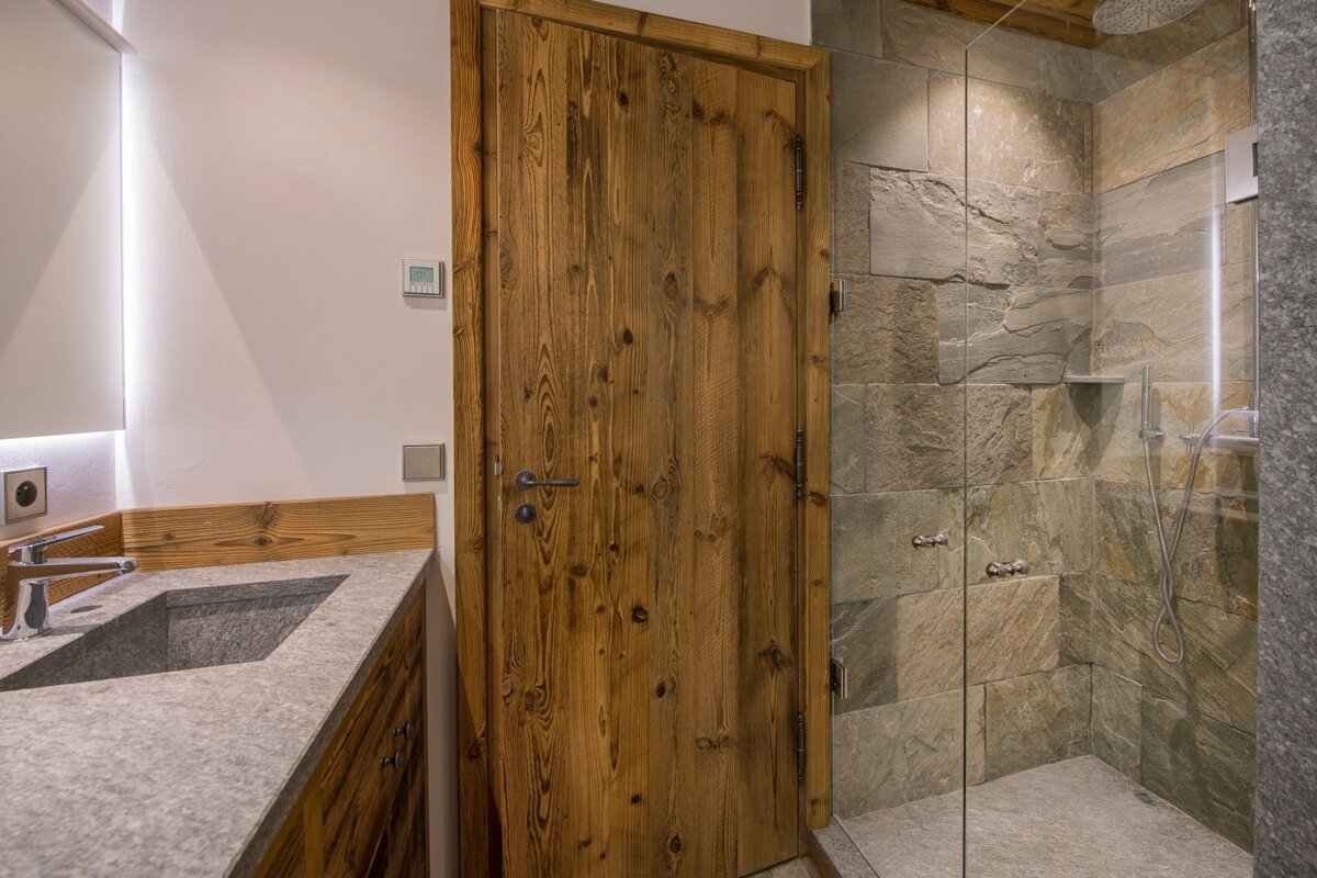 A stylish bathroom featuring a weathered wooden door, a sleek stone vanity, and a large shower with natural stone tiles and a glass enclosure.