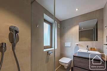 A modern bathroom featuring beige tiled walls, a glass shower partition, toilet, small window, and a wooden vanity with a white sink.