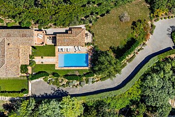 An aerial shot captures a charming villa with a bright blue pool, manicured green lawns, and red tiled roofs, surrounded by lush trees and a winding road.