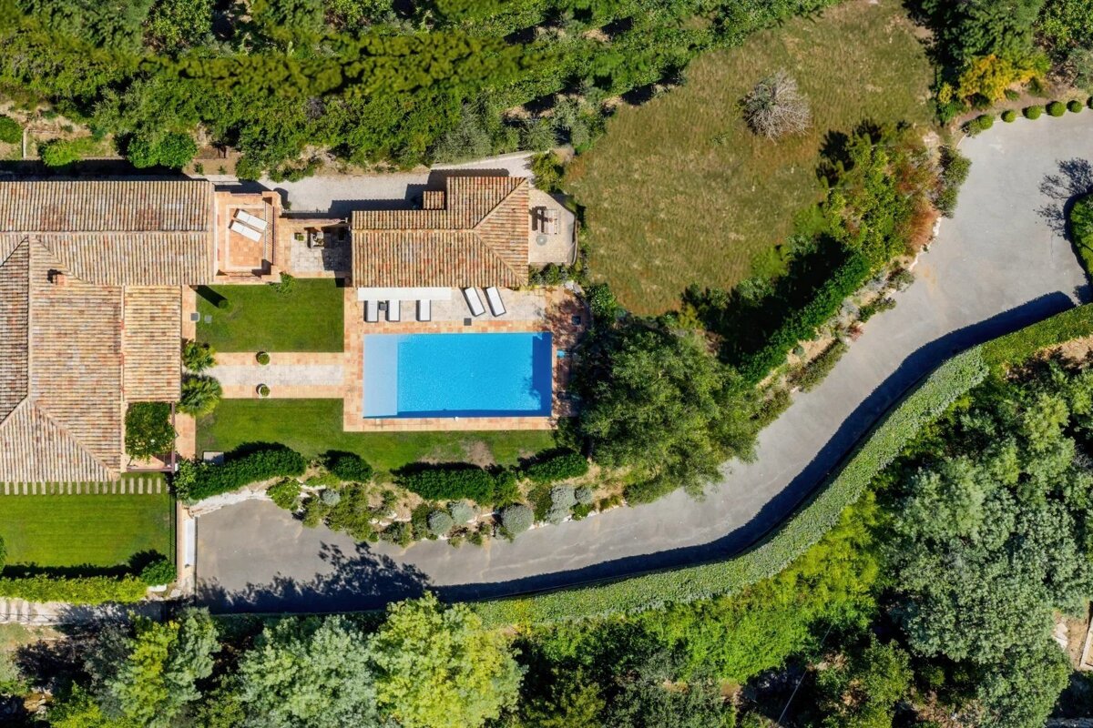 Aerial view of a villa with a bright blue pool, surrounded by lush green lawns, trees, and a winding driveway.