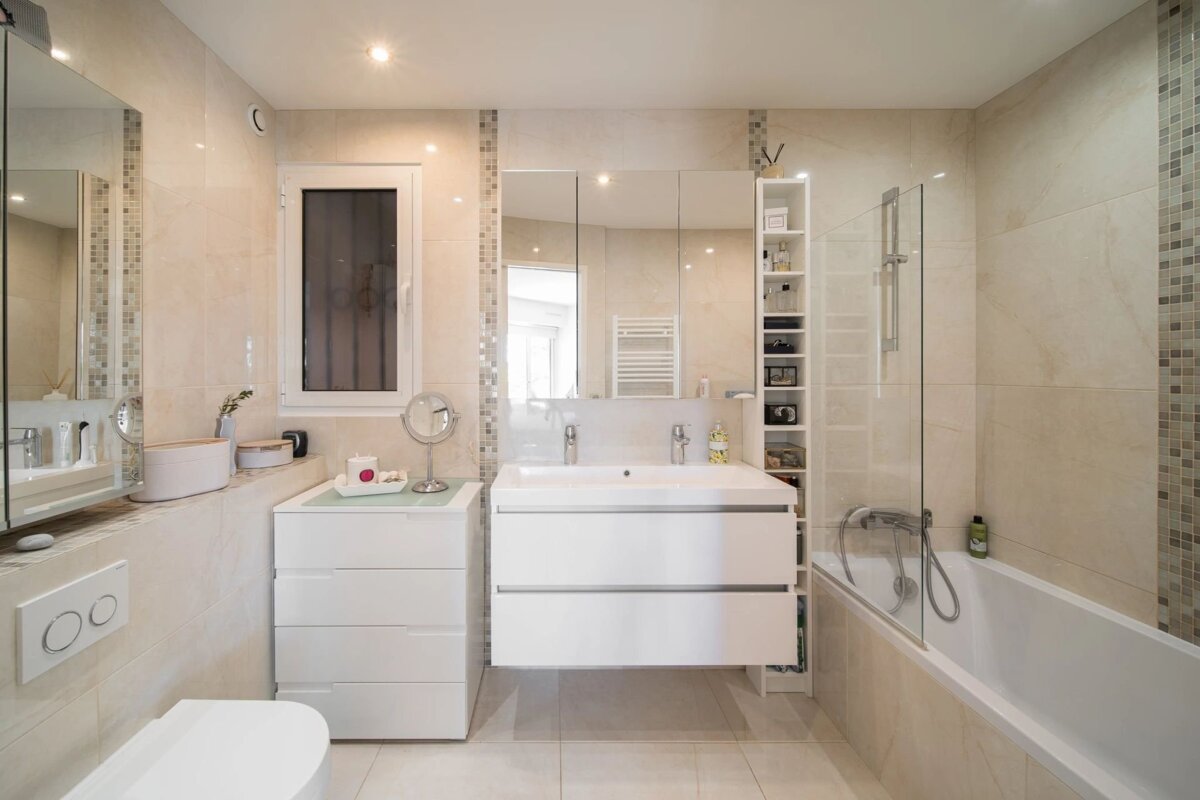 Modern bathroom with beige tiles, double sink vanity, large mirror cabinet, white toilet, bathtub with glass screen, and storage shelves.