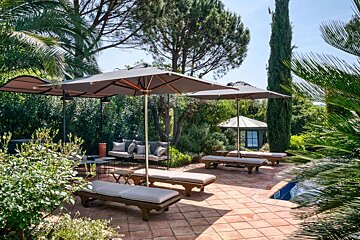 A sunny, lush outdoor patio with terracotta tiles, large umbrellas shading lounge chairs and a cozy seating area, surrounded by vibrant greenery and a small pool.
