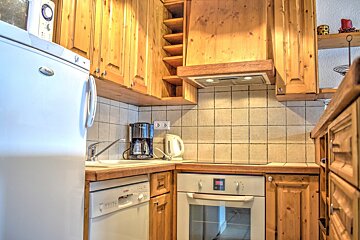 A compact kitchen with light wooden cabinets, white fridge, dishwasher, oven, tiled backsplash, coffee maker, and kettle.