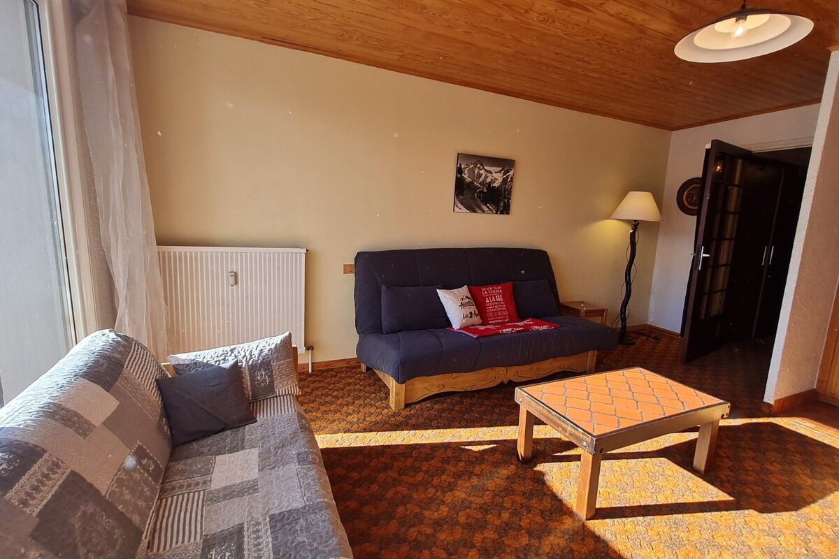 A cozy, sunlit living room with two sofas, a tiled coffee table, and a wooden ceiling. A floor lamp and a doorway are visible.