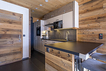 A cozy, modern kitchen with rustic wood paneling on the walls and ceiling. It features grey cabinets, dark countertops, and a breakfast bar.